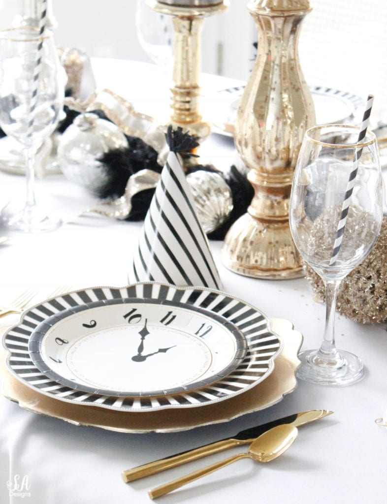 black white gold tablescape, black and white striped graces dinner plates, gold chargers, crystal chandelier, clock plates new years eve, new years eve game, new years eve food, mercury glass candlesticks, sugar paper la white black scalloped dinner napkins, party hat, mercury glass candlesticks, glitter candles. gold rimmed crystal goblets red wine glasses