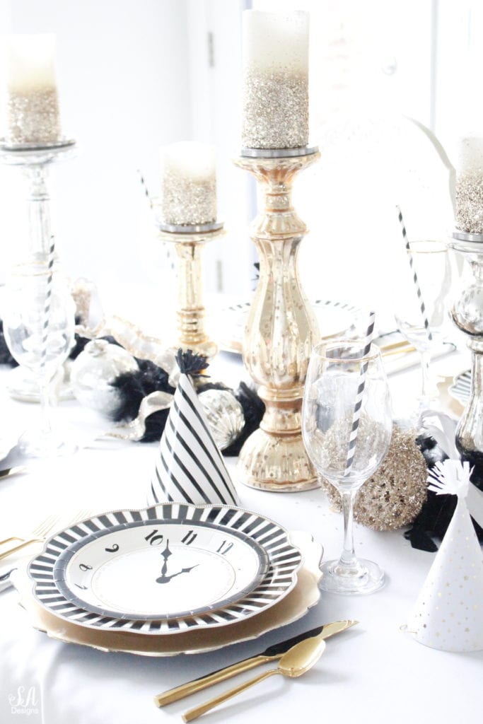 black white gold tablescape, black and white striped graces dinner plates, gold chargers, crystal chandelier, clock plates new years eve, new years eve game, new years eve food, mercury glass candlesticks, sugar paper la white black scalloped dinner napkins, party hat, mercury glass candlesticks, glitter candles. gold rimmed crystal goblets red wine glasses, gold silver ornaments, mercury glass ornaments with ribbon