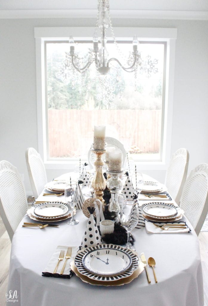 black white gold tablescape, black and white striped graces dinner plates, gold chargers, crystal chandelier, clock plates new years eve, new years eve game, new years eve food, mercury glass candlesticks, sugar paper la white black scalloped dinner napkins, party hat, mercury glass candlesticks, glitter candles