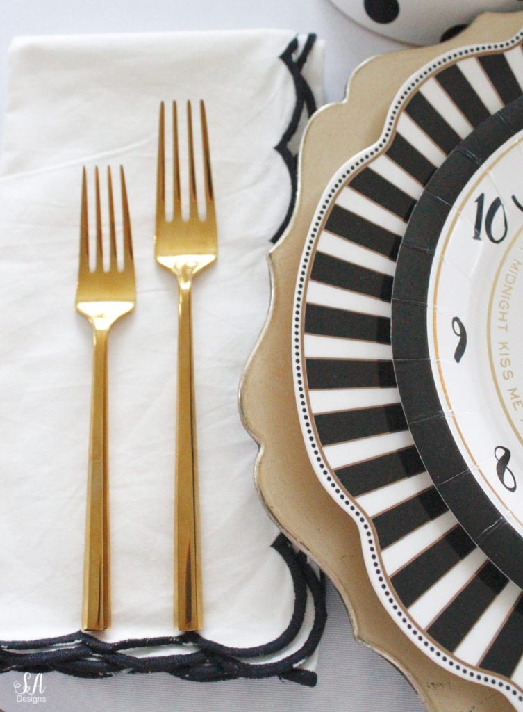 black white gold tablescape, black and white striped graces dinner plates, gold chargers, crystal chandelier, clock plates new years eve, new years eve game, new years eve food, mercury glass candlesticks, sugar paper la white black scalloped dinner napkins, party hat, mercury glass candlesticks, glitter candles