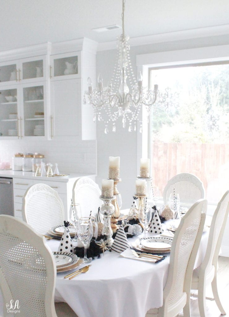 black white gold tablescape, black and white striped graces dinner plates, gold chargers, crystal chandelier, clock plates new years eve, new years eve game, new years eve food, mercury glass candlesticks, sugar paper la white black scalloped dinner napkins, party hat, mercury glass candlesticks, glitter candles