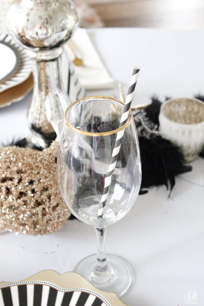 black white gold tablescape, black and white striped graces dinner plates, gold chargers, crystal chandelier, clock plates new years eve, new years eve game, new years eve food, mercury glass candlesticks, sugar paper la white black scalloped dinner napkins, party hat, mercury glass candlesticks, glitter candles. gold rimmed crystal goblets red wine glasses