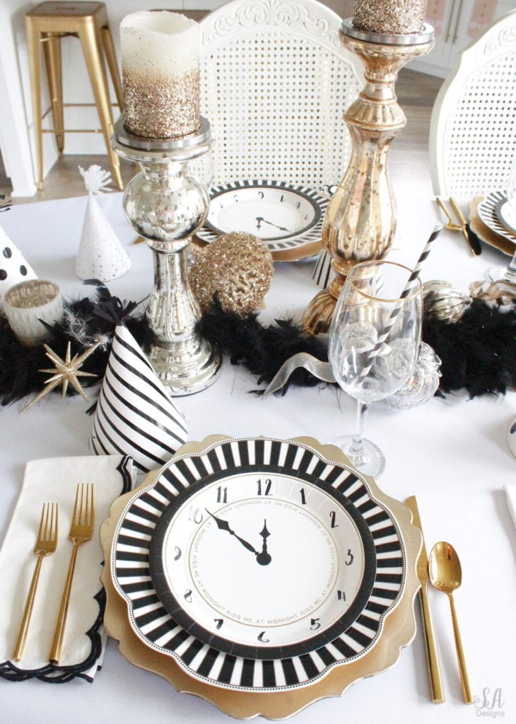 black white gold tablescape, black and white striped graces dinner plates, gold chargers, crystal chandelier, clock plates new years eve, new years eve game, new years eve food, mercury glass candlesticks, sugar paper la white black scalloped dinner napkins, party hat, mercury glass candlesticks, glitter candles