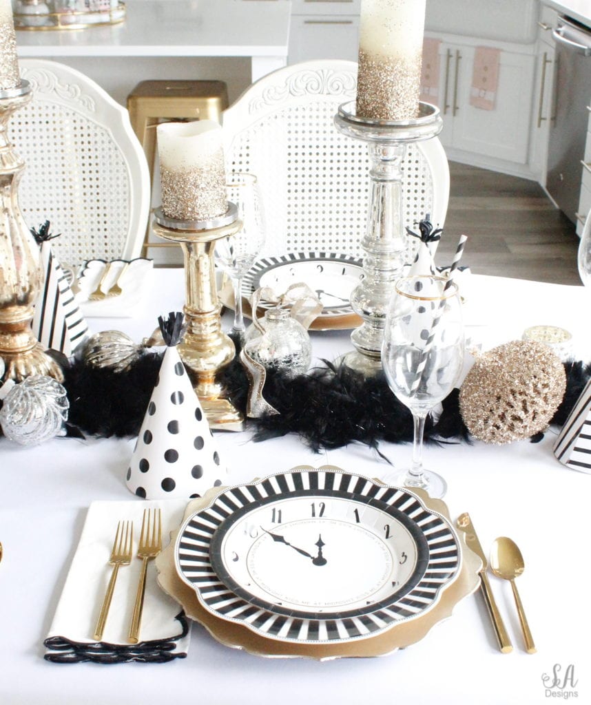 black white gold tablescape, black and white striped graces dinner plates, gold chargers, crystal chandelier, clock plates new years eve, new years eve game, new years eve food, mercury glass candlesticks, sugar paper la white black scalloped dinner napkins, party hat, mercury glass candlesticks, glitter candles. gold rimmed crystal goblets red wine glasses