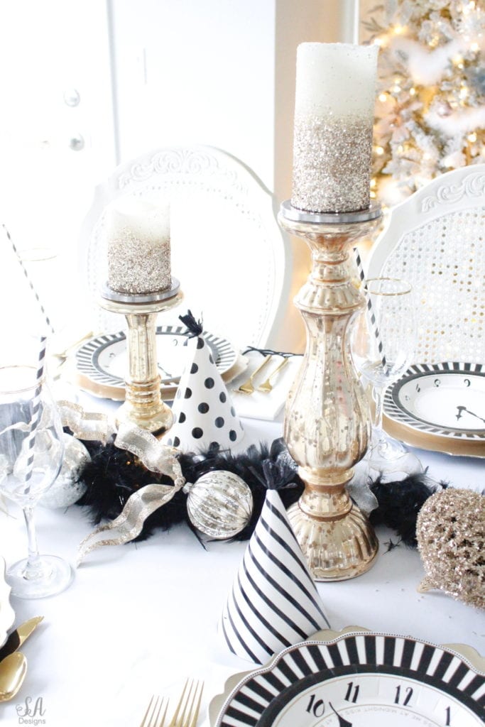 black white gold tablescape, black and white striped graces dinner plates, gold chargers, crystal chandelier, clock plates new years eve, new years eve game, new years eve food, mercury glass candlesticks, sugar paper la white black scalloped dinner napkins, party hat, mercury glass candlesticks, glitter candles. gold rimmed crystal goblets red wine glasses, gold silver ornaments, mercury glass ornaments with ribbon