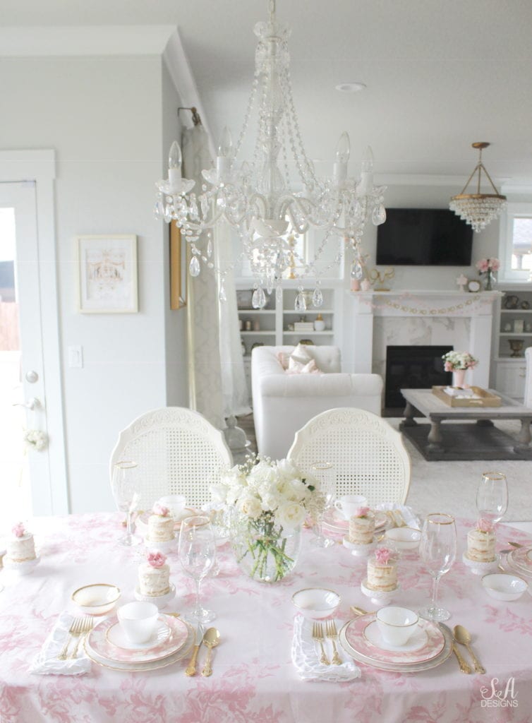 monique lhuillier pottery barn, pink floral tablecloth, pink floral plates, white kitchen design, vintage valentines day tablescape, valentines decor table, antique vintage milk glass plates teacups saucers gold rimmed, gold rimmed crystal goblets, vintage gold flatware, white ruffled napkins, individual mini naked cakes with fresh pink rose baby's breath