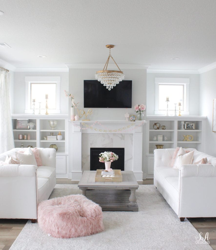 blush pink valentine's day decor, crystorama chandelier, white and gold interiors, white interiors, classy interiors, television above mantel mantle, tv above fireplace, valentines banner garland, white chesterfield tufted sofa, vintage pink milkglass, pink faux fur ottoman pouf, coffee table styling, interior design style stylish books for coffee table, fresh flowers