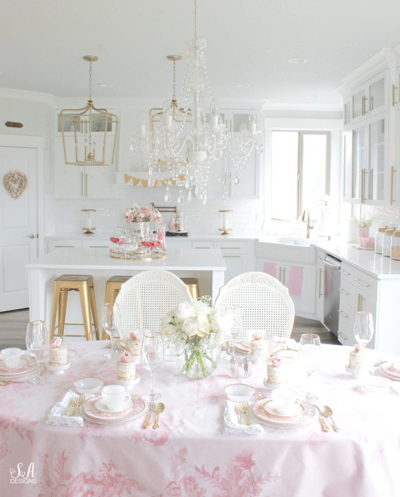 monique lhuillier pottery barn, pink floral tablecloth, pink floral plates, white kitchen design, vintage valentines day tablescape, valentines decor table, antique vintage milk glass plates teacups saucers gold rimmed, gold rimmed crystal goblets, vintage gold flatware, white ruffled napkins, individual mini naked cakes with fresh pink rose baby's breath