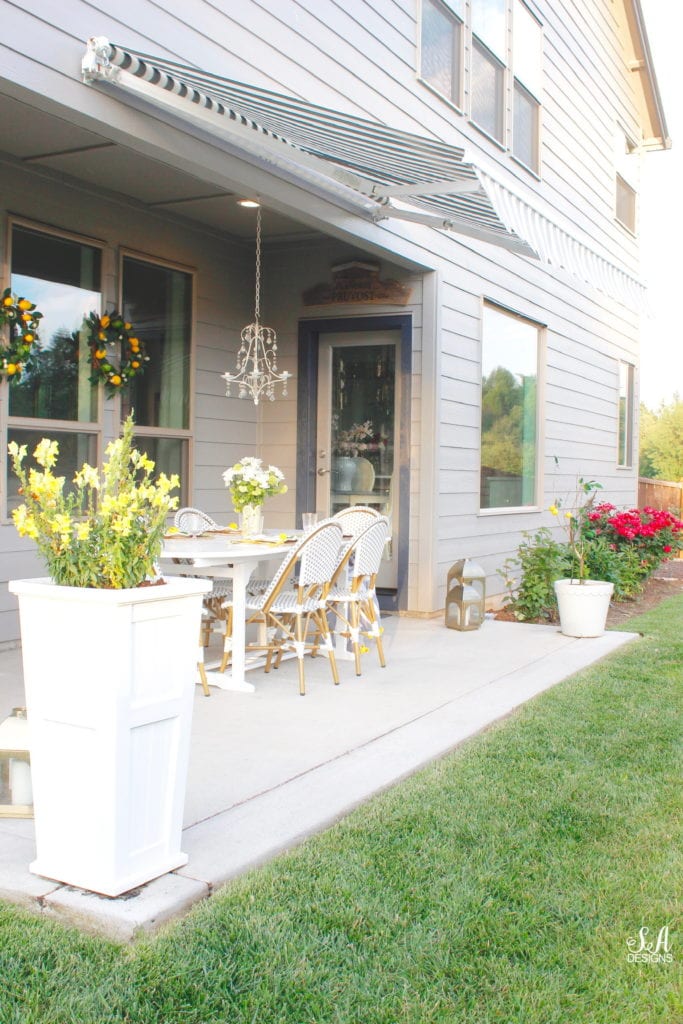 grey and white striped retractable awning, french outdoor patio, french style outdoor patio dining, french coastal backyard landscape dining outdoor design, english garden lemon tablescape, serena and lily outdoor umbrella, serena and lily grey white french bistro chairs, white oval outdoor dining patio table, outdoor patio table dining set, gold brass bar cart, lemon and fruit floral arrangement, outdoor patio chandelier, lemon tree