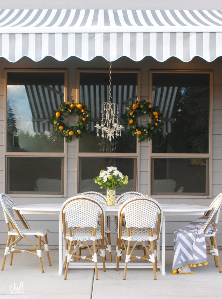 grey and white striped retractable awning, french outdoor patio, french style outdoor patio dining, french coastal backyard landscape dining outdoor design, english garden lemon tablescape, serena and lily outdoor umbrella, serena and lily grey white french bistro chairs, white oval outdoor dining patio table, outdoor patio table dining set, gold brass bar cart, lemon and fruit floral arrangement, outdoor patio chandelier, lemon tree