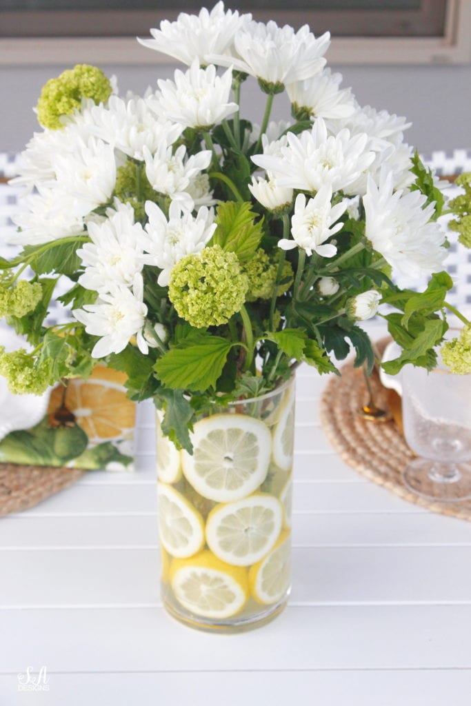 fruit and floral arrangement, lemon and flower arrangement, flower arrangement with lemon slices in vase,grey and white striped retractable awning, french outdoor patio, french style outdoor patio dining, french coastal backyard landscape dining outdoor design, english garden lemon tablescape, serena and lily outdoor umbrella, serena and lily grey white french bistro chairs, white oval outdoor dining patio table, outdoor patio table dining set, gold brass bar cart, lemon and fruit floral arrangement, outdoor patio chandelier, lemon tree