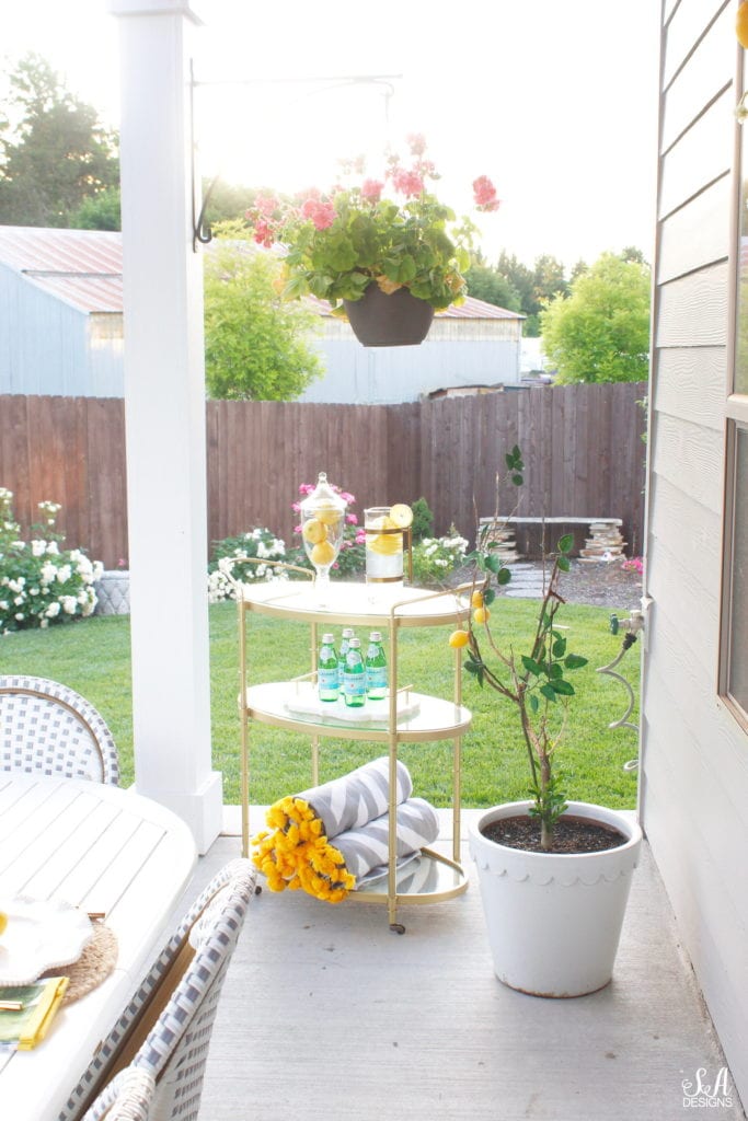 grey and white striped retractable awning, french outdoor patio, french style outdoor patio dining, french coastal backyard landscape dining outdoor design, english garden lemon tablescape, serena and lily outdoor umbrella, serena and lily grey white french bistro chairs, white oval outdoor dining patio table, outdoor patio table dining set, gold brass bar cart, lemon and fruit floral arrangement, outdoor patio chandelier, lemon tree