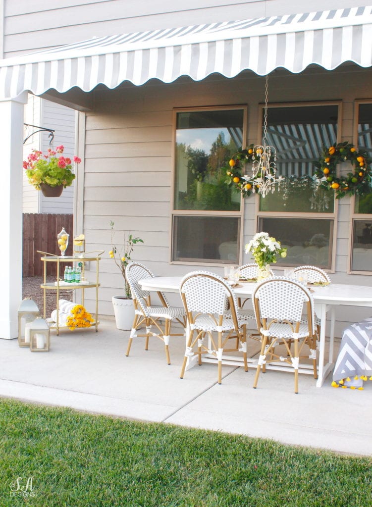 grey and white striped retractable awning, french outdoor patio, french style outdoor patio dining, french coastal backyard landscape dining outdoor design, english garden lemon tablescape, serena and lily outdoor umbrella, serena and lily grey white french bistro chairs, white oval outdoor dining patio table, outdoor patio table dining set, gold brass bar cart, lemon and fruit floral arrangement, outdoor patio chandelier, lemon tree