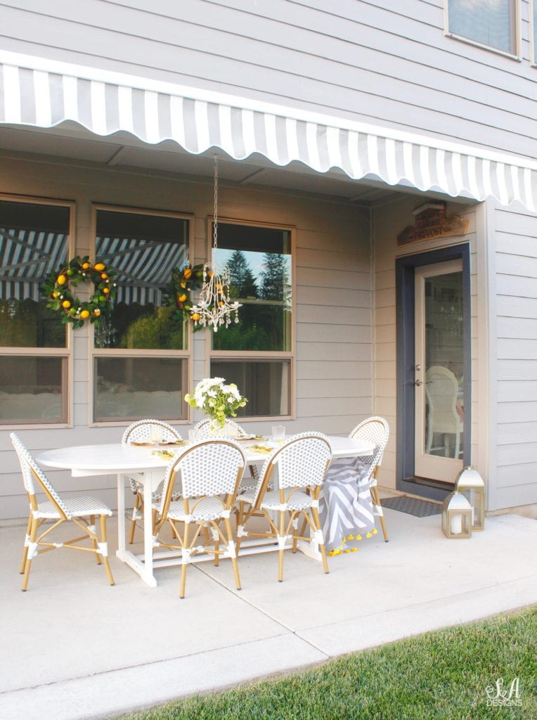 grey and white striped retractable awning, french outdoor patio, french style outdoor patio dining, french coastal backyard landscape dining outdoor design, english garden lemon tablescape, serena and lily outdoor umbrella, serena and lily grey white french bistro chairs, white oval outdoor dining patio table, outdoor patio table dining set, gold brass bar cart, lemon and fruit floral arrangement, outdoor patio chandelier, lemon tree