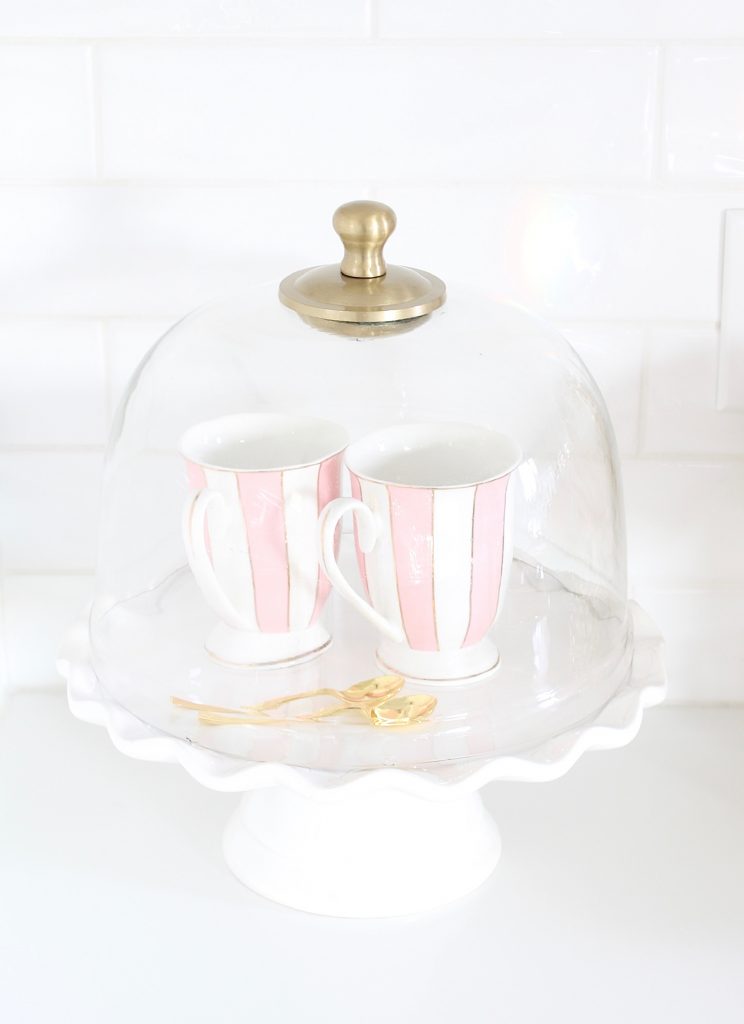 striped pink teacups stacked on white ruffle cake stand under cloche dome, pink and blue spring kitchen decor, white kitchen design, white and gold kitchen, voluspa large 5 wick candle, voluspa macaron candles, white hood, white and gold kitchen, kitchen goals, Caitlin Wilson blue bread cutting board French blue, blogger kitchen inspiration