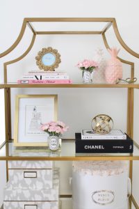 white and gold office, blush office, glam office, etagere gold shelves, Maltese dog, nesting boxes, home office, feminine office, diptyque candles used a flower vase, upcycled diptyque candle holder, white ginger jar pink cherry blossom stems, white matted gold frame set, office accessories, desk accessories, leopard slippers, white and gold leather office chair with wheels, pink pom pom throw blanket, blush tassel tote, gold office shelves, office shelving, how to decorate shelves, blogger office